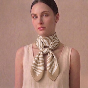 Peut inclure: Un foulard en soie à rayures zébrées beige et blanc noué autour du cou. Le foulard est de forme carrée avec un motif répété de rayures beiges et blanches. Le foulard est porté sur un haut de couleur claire.