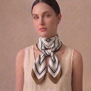 Peut inclure: Un foulard en soie avec un motif géométrique blanc, noir et marron. Le foulard est noué autour du cou, les extrémités pendantes. Il est de forme carrée et fabriqué dans un tissu doux et fluide.