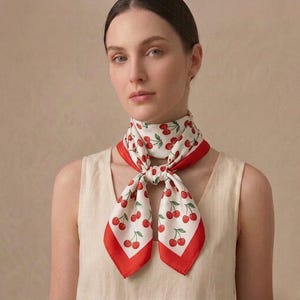Peut inclure: Foulard blanc orné de motifs de cerises rouges et vertes, bordé de rouge. Le foulard est noué autour du cou.