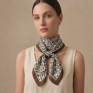 Könnte beinhalten: Ein Seidenschal mit Leopardenmuster in Schwarz und Creme, umrandet von einem braunen Rand. Der Schal ist um den Hals geknotet und bildet ein stilvolles Accessoire.