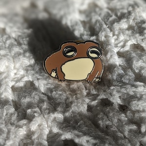 May include: A brown and cream frog enamel pin with a gold border. The pin is set against a textured, off-white background. The frog has a cartoon design with a relaxed expression.