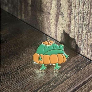 May include: A green frog pin wearing an orange pumpkin costume. The pin has a gold outline and is set against a wooden background. The frog's eyes are small and black, and the pumpkin has a stem.