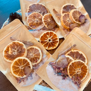 May include: Four clear bags filled with bath salts, dried orange slices, cinnamon sticks, and dried rose petals. The bags are made of clear plastic and have a brown paper backing.