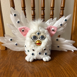 May include: A white plush toy with multiple eyes and pink ears, adorned with white feather wings, and set on a wooden chair. The toy has a beak and a tuft of fur on its head. The wings are decorated with blue and black eyes.