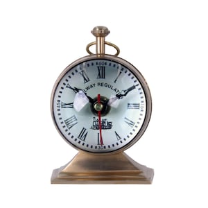 Puede incluir: Un reloj Railway Regulator redondo de color latón con esfera blanca y números romanos negros. El reloj tiene manecillas negras y un segundero rojo. La base es cuadrada y el reloj tiene un pequeño lazo en la parte superior.