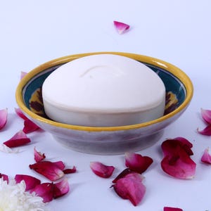 Puede incluir: Una pastilla de jabón redonda y blanca descansa en un jabonera de cerámica decorativa. El plato tiene un borde amarillo, un interior turquesa con diseños florales y una base lavanda. Pétalos de rosa rosas y rojos están esparcidos.