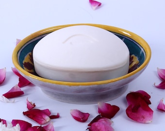 May include: A round, white bar of soap sits in a decorative ceramic soap dish. The dish has a yellow rim, a teal interior with floral designs, and a lavender base. Scattered around are pink and red rose petals.