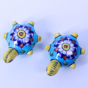 Könnte beinhalten: Zwei Keramik-Schildkrötenfiguren. Die Schildkröten haben einen hellblauen Panzer mit einem Blumendesign in Weiß, Blau und Braun. Kopf und Füße sind gelb. Die Schildkröten sind dekorativ.