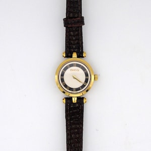 Könnte beinhalten: Eine goldfarbene Armbanduhr mit weißem Zifferblatt und schwarzen römischen Ziffern. Die Uhr hat ein braunes Lederarmband mit strukturiertem Muster. Auf dem Zifferblatt steht der Markenname "GUCCI".