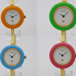 Puede incluir: Cuatro relojes Gucci con correas de diferentes colores. Los relojes tienen esferas blancas con agujas negras y un logotipo Gucci dorado. Las correas son naranja, verde, azul y rosa.