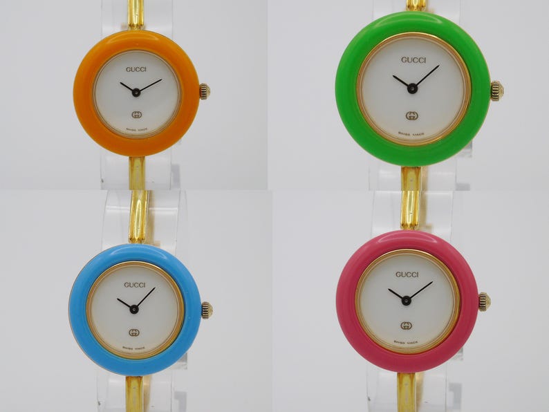 Puede incluir: Cuatro relojes Gucci con correas de diferentes colores. Los relojes tienen esferas blancas con agujas negras y un logotipo Gucci dorado. Las correas son naranja, verde, azul y rosa.