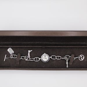 Può includere: Un bracciale a catena argentata con quadrante e ciondoli decorativi, tra cui una targhetta per cani, uno stivale e una staffa. Il bracciale è esposto in una scatola marrone con interno scuro.