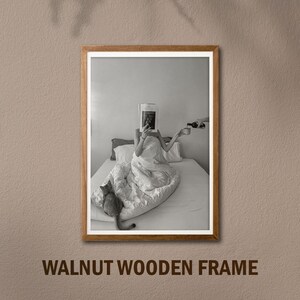 Op de afbeelding: Een zwart-witfoto van een persoon in bed die een tijdschrift leest. De persoon is bedekt met een wit deken en heeft een kat naast zich zitten. De foto is ingelijst in een walnoot houten lijst.