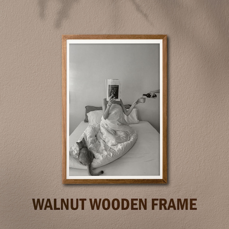 Op de afbeelding: Een zwart-witfoto van een persoon in bed die een tijdschrift leest. De persoon is bedekt met een wit deken en heeft een kat naast zich zitten. De foto is ingelijst in een walnoot houten lijst.