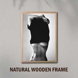 May include: Black and white photo of a person pulling a black shirt off their body. The photo is framed in a natural wood frame.
