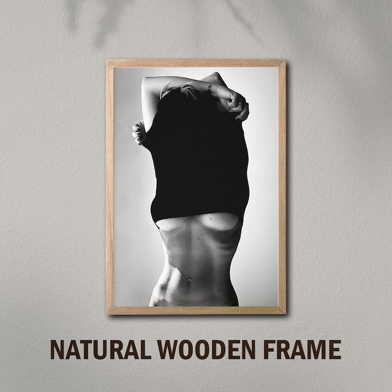 May include: Black and white photo of a person pulling a black shirt off their body. The photo is framed in a natural wood frame.