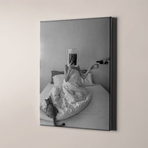 Op de afbeelding: Een zwart-witfoto van een persoon in bed, die een tijdschrift leest met hun gezicht bedekt. De persoon houdt een kopje in de ene hand en een fles wijn in de andere. Een kat zit op het bed op de voorgrond.