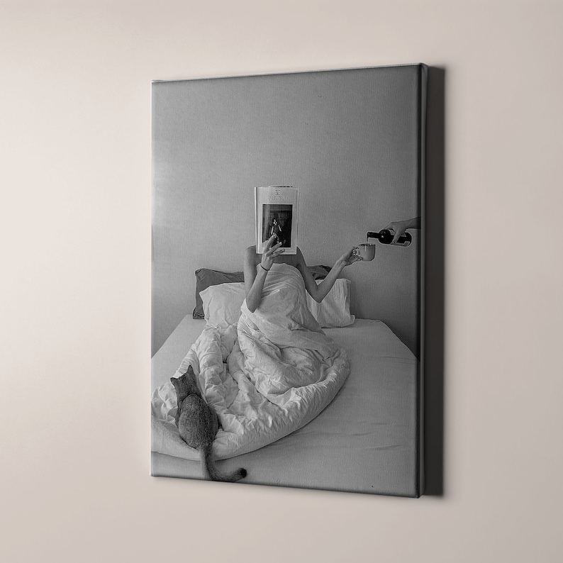 Op de afbeelding: Een zwart-witfoto van een persoon in bed, die een tijdschrift leest met hun gezicht bedekt. De persoon houdt een kopje in de ene hand en een fles wijn in de andere. Een kat zit op het bed op de voorgrond.