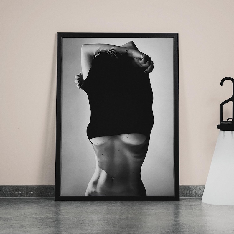 May include: Black and white photograph of a person pulling a black shirt off their shoulders, revealing their torso.