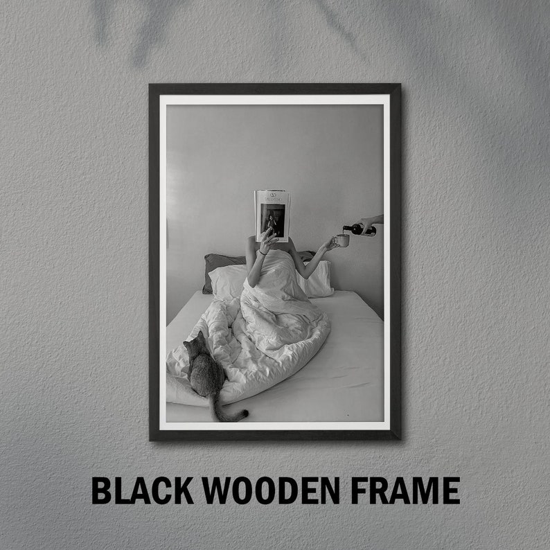 Op de afbeelding: Zwart-witfoto van een persoon in bed met een tijdschrift over hun gezicht, die een kopje vasthoudt. Een kat zit op het bed. De foto is ingelijst in een zwart houten frame.