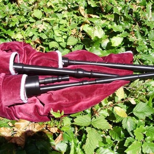 May include: A set of three black bagpipes with a red velvet bag and white trim. The bagpipes are lying on a bed of green leaves.