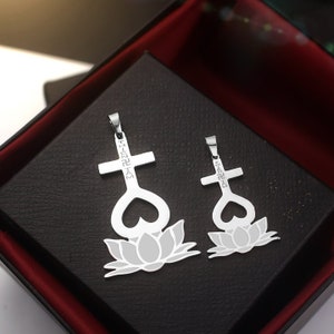 May include: Two silver pendants, one larger than the other, feature a cross with a heart and a lotus flower design. The pendants are on a black background.