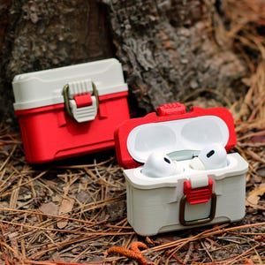 Puede incluir: Dos estuches protectores rojos y blanquecinos para auriculares inalámbricos. Un estuche está abierto, revelando auriculares blancos en su interior. Los estuches tienen un cierre de pestillo y están diseñados para parecerse a pequeñas cajas de aparejos. Los estuches descansan sobre un lecho de agujas de pino.