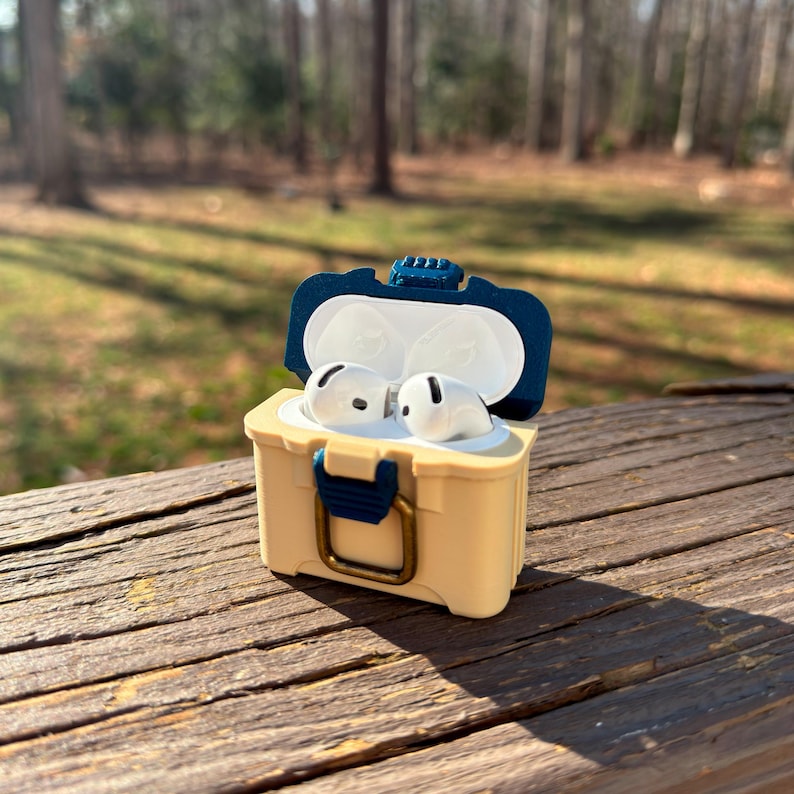 Puede incluir: Una funda protectora beige y azul marino para auriculares inal&aacute;mbricos, que se asemeja a un peque&ntilde;o cofre, est&aacute; abierta para revelar auriculares blancos. La funda tiene un cierre y detalles en azul marino, y est&aacute; sobre una superficie de madera.