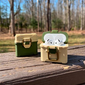 May include: A green and beige protective case for AirPods with a latch closure. The case is open and shows the white AirPods inside.
