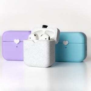 Pu&ograve; includere: Tre custodie per AirPod, una blu chiaro, una viola e una bianca con un motivo maculato. La custodia bianca &egrave; aperta, rivelando due auricolari AirPod bianchi.