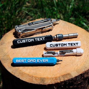 以下が含まれることがあります： カスタムテキストの彫刻が施されたマルチツールが4つあります。ツールは、シルバーのマルチツール、黒いマルチツールに「CUSTOM TEXT」、白いマルチツールに「CUSTOM TEXT」、青いマルチツールに「BEST DAD EVER」と刻印されています。