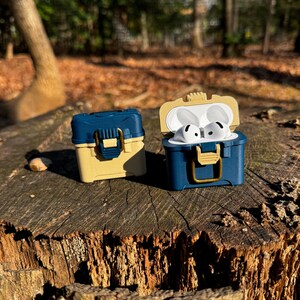 Puede incluir: Dos estuches en miniatura azules y beige con forma de caja de aparejos de pesca, uno abierto con auriculares blancos en su interior. Los estuches est&aacute;n sobre un toc&oacute;n de &aacute;rbol en un entorno forestal. Los estuches tienen una tapa con bisagras y un cierre dorado.