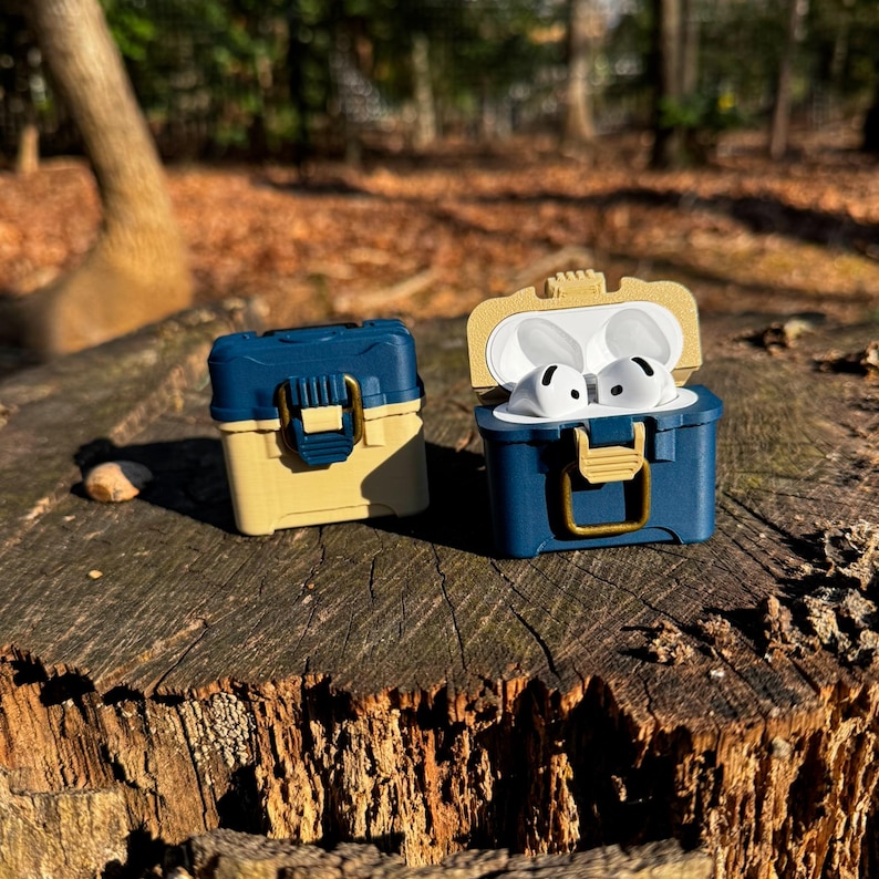 Puede incluir: Dos estuches en miniatura azules y beige con forma de caja de aparejos de pesca, uno abierto con auriculares blancos en su interior. Los estuches est&aacute;n sobre un toc&oacute;n de &aacute;rbol en un entorno forestal. Los estuches tienen una tapa con bisagras y un cierre dorado.
