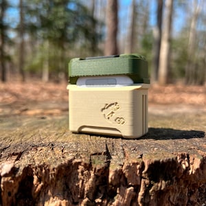 May include: A beige and green protective case for a small electronic device. The case has a textured surface and a logo on the front. The case is sitting on a tree stump.