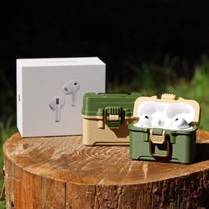 May include: A white box with a pair of white earbuds and two green and tan cases for earbuds are displayed on a tree stump. One case is open, revealing white earbuds inside. The cases have a latch closure.
