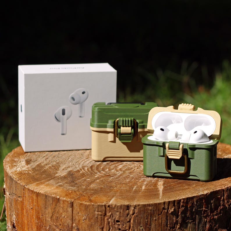 May include: A white box with a pair of white earbuds and two green and tan cases for earbuds are displayed on a tree stump. One case is open, revealing white earbuds inside. The cases have a latch closure.