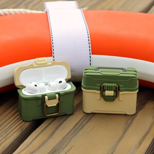 May include: Two green and tan AirPod cases, one open with white earbuds inside. The cases have a latch closure and a handle on top. The background includes an orange life preserver and a wooden surface.