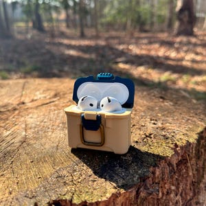 Puede incluir: Una funda protectora beige y azul marino para auriculares inal&aacute;mbricos, con forma de peque&ntilde;a nevera, est&aacute; abierta sobre un toc&oacute;n de &aacute;rbol. En el interior, se ven dos auriculares blancos. La funda tiene un cierre azul y una tapa azul.