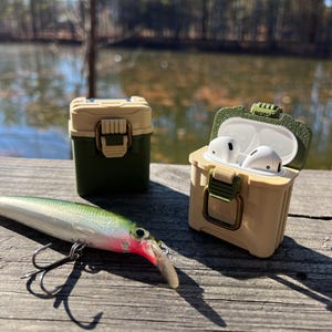 May include: A green and beige tackle box style case for Airpods with a fishing lure in the foreground. The case is open and the Airpods are inside.