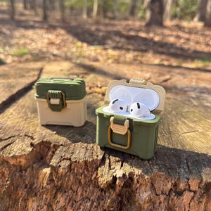 May include: Two green and beige protective cases for wireless earbuds. The cases are designed to look like miniature toolboxes with latches and handles. One case is open, revealing two white earbuds inside.