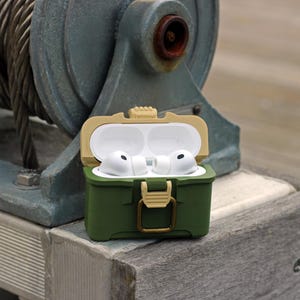 May include: An open, olive green case with a tan lid, holding white wireless earbuds. The case has a gold-colored clasp and is sitting on a weathered wooden surface. The background includes a large, industrial-looking metal structure.