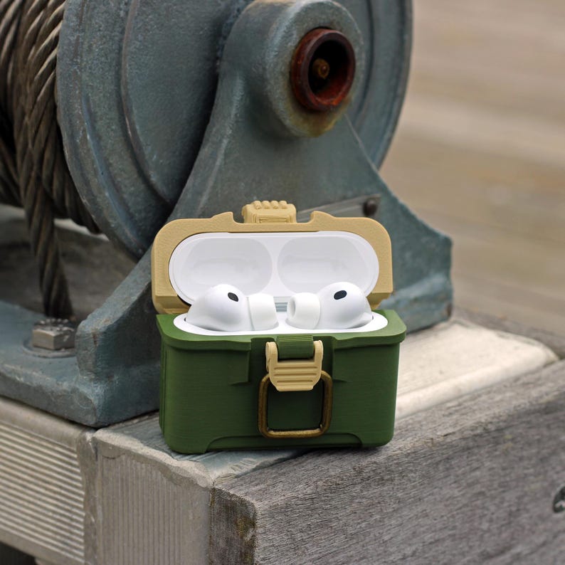 May include: An open, olive green case with a tan lid, holding white wireless earbuds. The case has a gold-colored clasp and is sitting on a weathered wooden surface. The background includes a large, industrial-looking metal structure.