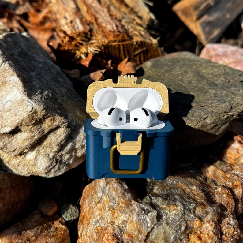Puede incluir: Auriculares inal&aacute;mbricos blancos dentro de un estuche azul marino y dorado, con forma de cofre del tesoro. El estuche est&aacute; abierto, revelando los auriculares. El fondo presenta rocas y follaje.