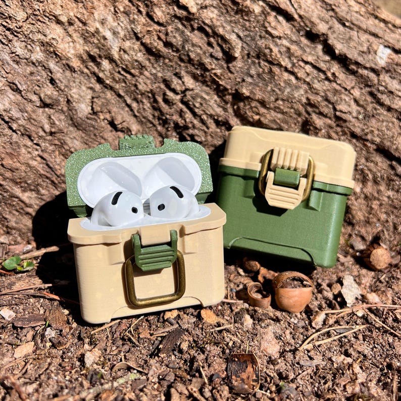 May include: Two green and beige plastic cases with a latch closure. One case is open and contains two white wireless earbuds. The cases are on a brown and green background.