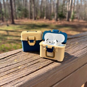 Puede incluir: Dos peque&ntilde;as cajas beige y azules, que parecen cofres del tesoro en miniatura, se exhiben al aire libre. Una caja est&aacute; cerrada, mientras que la otra est&aacute; abierta, revelando auriculares blancos. Las cajas tienen cierres y bisagras doradas, con un acento azul en la tapa.