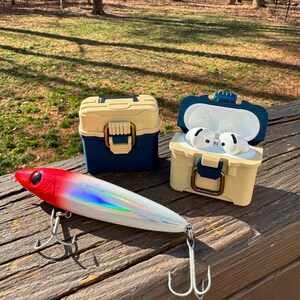 Puede incluir: Un se&ntilde;uelo de pesca rojo y blanco con detalles plateados y tres anzuelos descansa sobre una superficie de madera junto a dos peque&ntilde;as cajas en forma de caja. Una caja es azul y beige, la otra es beige con la parte superior azul y contiene auriculares blancos.