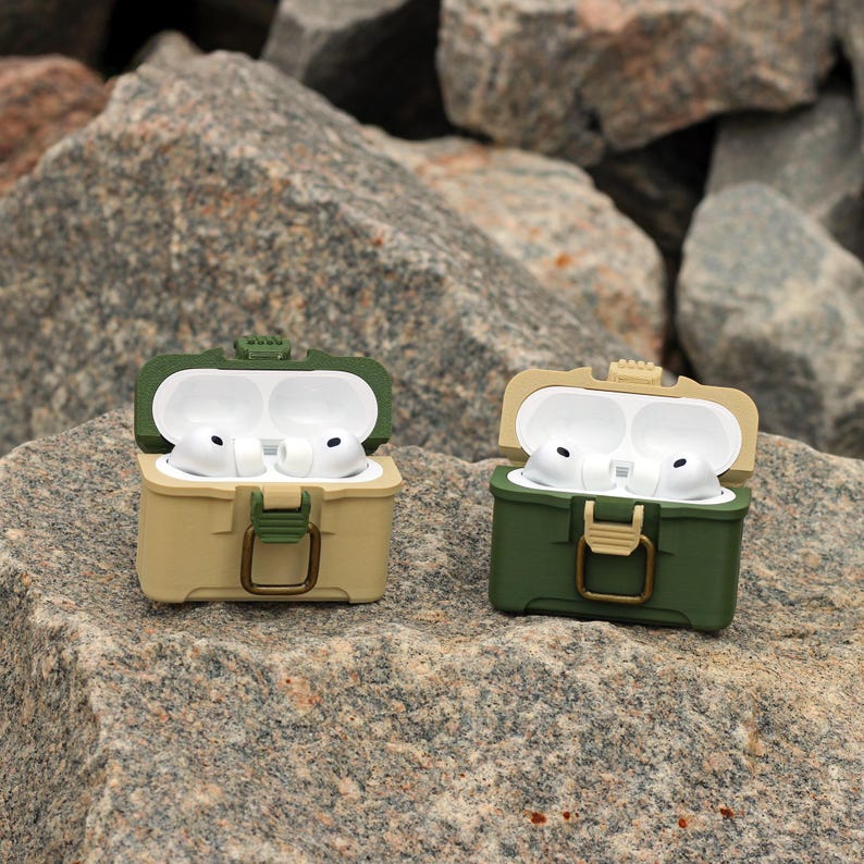 May include: Two open, olive green and tan AirPod cases with white AirPods inside. The cases have a hinged lid and a clasp closure. The cases are sitting on a large, gray rock.