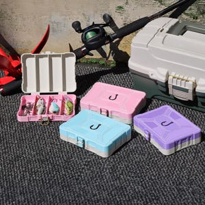 Puede incluir: Una colección de cajas de aparejos de pesca en varios colores, incluyendo rosa, azul y morado. Cada caja tiene un símbolo de anzuelo y un cierre. La imagen también muestra una caña de pescar, una caja blanca y una caja rosa con señuelos.