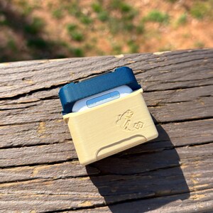 Puede incluir: Una funda protectora beige y azul marino para auriculares inal&aacute;mbricos. La funda tiene una tapa con bisagras y un peque&ntilde;o gr&aacute;fico de pez en la parte delantera. Los auriculares son blancos.