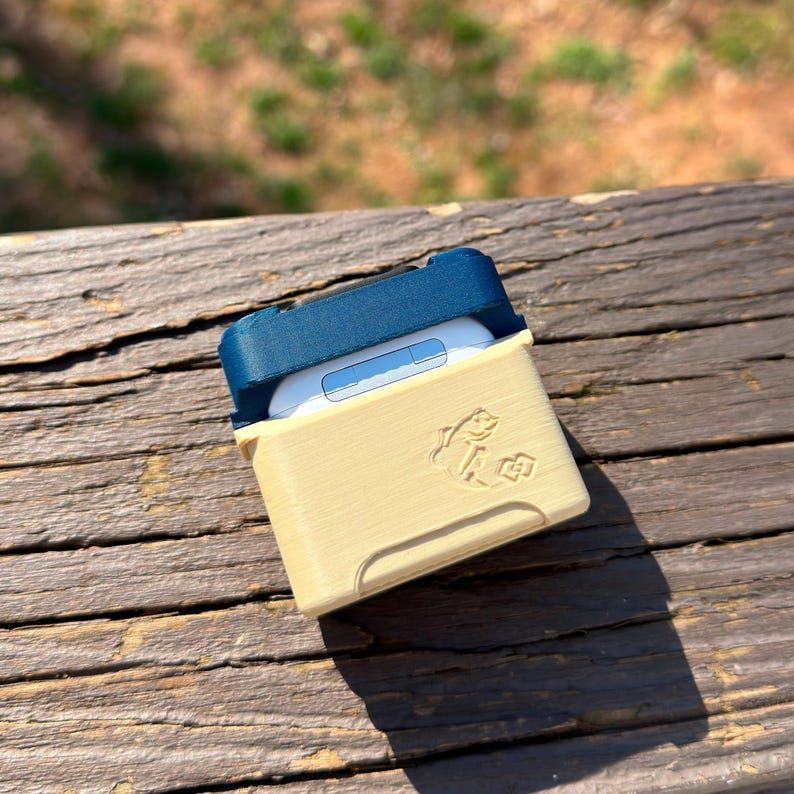 Puede incluir: Una funda protectora beige y azul marino para auriculares inal&aacute;mbricos. La funda tiene una tapa con bisagras y un peque&ntilde;o gr&aacute;fico de pez en la parte delantera. Los auriculares son blancos.